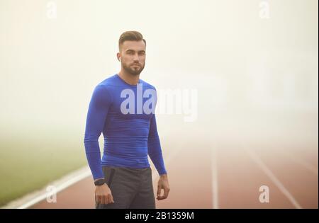 Porträt eines jungen glücklichen männlichen Athleten in Kopfhörern und einer Uhr in einem Stadion im Nebel. Stockfoto