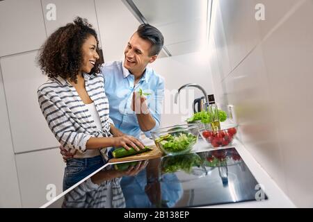 Romantisches Datum. Junges, multiethnisches Paar, das beim Küchen-Dinner steht und Salat umarmte, sich gegenseitig lachend zu betrachten Stockfoto