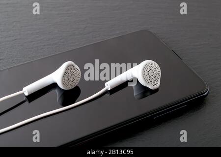 Weiße Ohrhörer spiegeln sich in einem schwarzen Smartphone-Bildschirm auf einer schwarzen Steinoberfläche wider. Modernes, persönliches Gerät mit Touchscreen für Musik und Kommunikation. Stockfoto