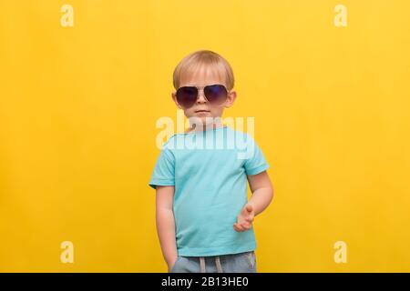 Kinderporträt eines 3 Jahre alten blonden Jungen in einem blauen T-Shirt und einer Sonnenbrille auf hellgelbem Frühlingsommer-Hintergrund mit Platz für Text. Anziehen Stockfoto