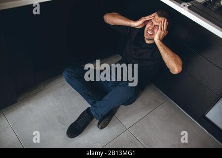 Der junge Geschäftsmann sitzt auf dem Boden und in der Küche und hält die Hände am Kopf. Kopfschmerzen und starke schreckliche Schmerzen. Guy leidet unter einer Störung oder einem phychologischen Probel Stockfoto