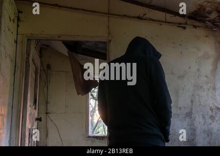 Blick auf den Rücken einer geheimnisvollen Kapuzenfigur in einem zerstörten, verlassenen Haus. Dunkle, stimmungsvolle Bearbeitung. Stockfoto