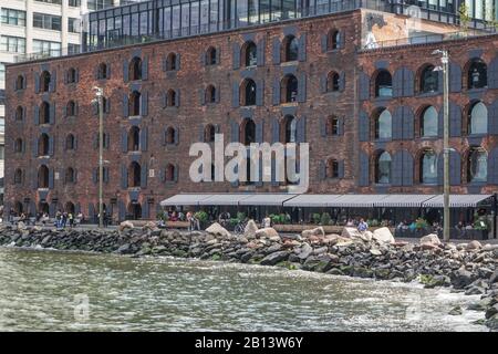 Brooklyn, New York: Menschen, die sich in einem Café in einem umgebauten Lagerhaus an der Küste von Brooklyn entspannen, während der East River gegen Felsen am Ufer ladet. Stockfoto