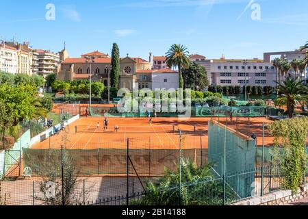 Overhead-Blick als Erwachsene trainieren Kinder beim Tennisspiel auf zwei Schmutzplätzen mit der Stadt Menton France dahinter, auf dem französischen Rivera. Stockfoto