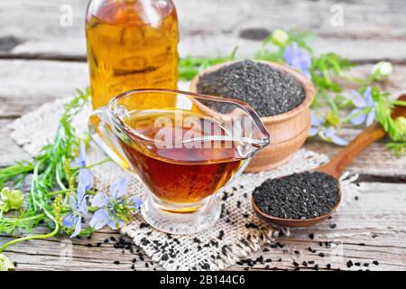 Nigella sativa Öl in Sauceboot und Flasche, Samen in einem Löffel und schwarzes Kümmelmehl in einer Schüssel auf dem Burlap, Kalingi-Zweige mit blauen Blumen und grünem Leav Stockfoto