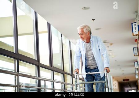 Älterer Aisian Mann, der mit Mühe einen Wanderer im Flur des Pflegeheims benutzt Stockfoto