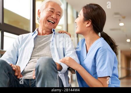 Junge freundliche asiatische Pflegekräfte unterhalten sich im Flur des Pflegeheims mit einem glücklichen Senioren-Mann Stockfoto