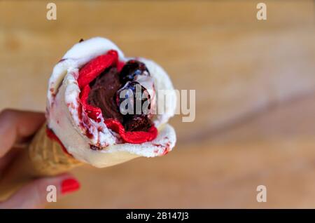 Frau, die ein rosenförmiges Eis mit Amarena, Schokolade und Erdbeeraroma hält, mit einer Kirsche oben in einem Zuckerkegel in Nizza, Frankreich Stockfoto