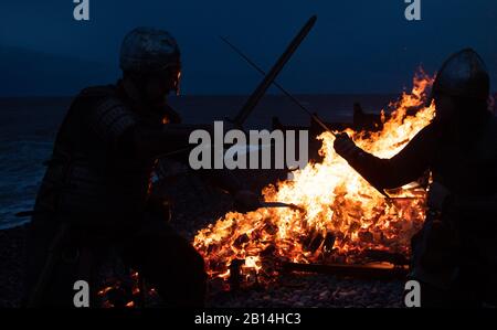 Reenactor im Kampf, während ein Langboot am Strand in Sheringham in Norfolk während des Sheringham Viking Festivals brennt, einer Feier des Norse Erbes der Küstenstadt. Stockfoto