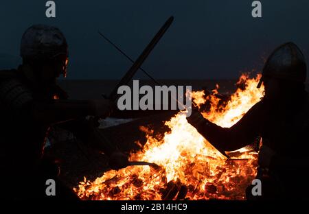 Reenactor im Kampf, während ein Langboot am Strand in Sheringham in Norfolk während des Sheringham Viking Festivals brennt, einer Feier des Norse Erbes der Küstenstadt. Stockfoto