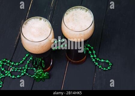 Zwei Gläser dunkles Bier und traditionelles Kleeblattdekor auf schwarzem Holztisch. Festliche Komposition des St. Patric'S Day Stockfoto