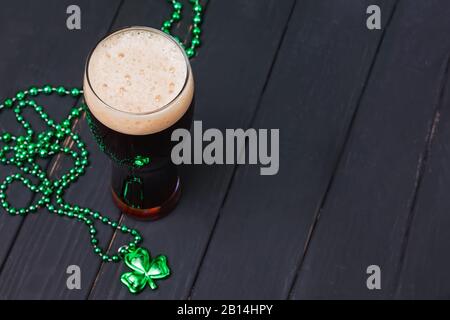 Glas dunkles Bier und traditionelles Kleeblattdekor auf schwarzem Holztisch. Festliche Komposition des St. Patric'S Day Stockfoto