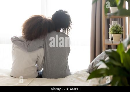 Liebevolle afrikanische alleinerziehende Mutter umarmen Teenager-Tochter, die auf dem Bett sitzt Stockfoto