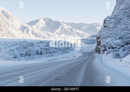 Winterlich verschneite Landschaft eines Bergtals mit einem Fluss mit einer rutschigen eisigen Straße mit steilen gefährlichen Kurven und Straßenschildern Stockfoto