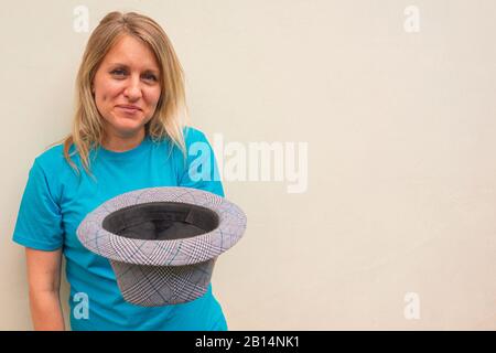 Lächelnde Frau hält einen Hut und bittet um Hilfe. Weißes Mädchen bleibt und fleht Geld oder Panhandling an. Ironie-Konzept. Vorderportrait mit fröhlichen Gesichts. Kopierspeicherplatz ist Stockfoto