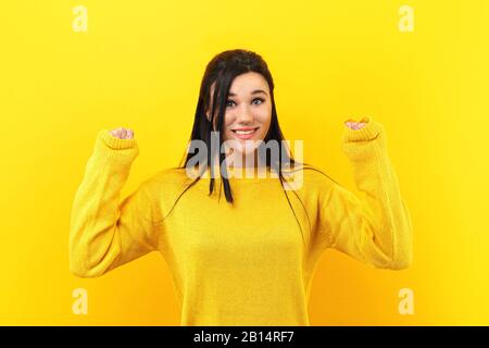 Brünette Mädchen ihren Erfolg und Sieg ballte die Fäuste mit Freude Freude. Glücklichen Frau in einem gelben Pullover in die Kamera lächeln Stockfoto