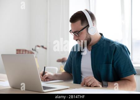 Fokussierter junger Mann, der das Webinar zum Thema Bildung beobachtet und Notizen aufschreibt. Stockfoto