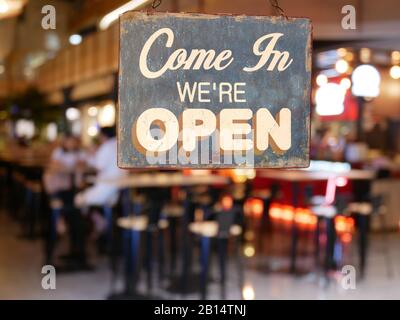 Ein Business Vintage Schild, das auf dem Cafe/Restaurant Fenster steht: „Come in We're Open“. Bild von abstrakten Unschärfe Restaurant mit Menschen. Stockfoto