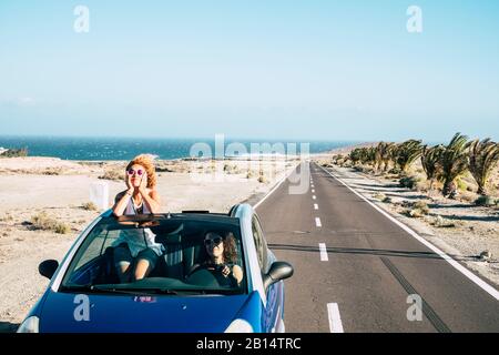 Reisefreunde und Transport mit blauem Cabrio-Auto und ein paar Erwachsene Frauen haben Spaß zusammen auf einer langen Straße mit Ozean in Backgr zu fahren Stockfoto