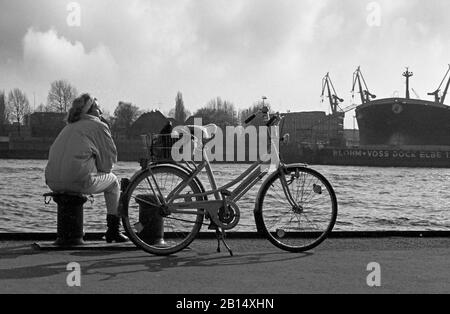 Lady sitzt mit ihrem Fahrrad auf der Quayside auf St. Pauli Landungsbrücken und blickt über die Elbe zum Schiffbaukomplex von Blohm & Voss, Hamburg, Deutschland, um 1988. Schwarzweiß-Filmfoto Stockfoto