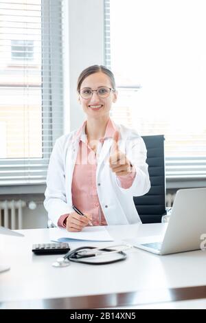 Arzt Frau geben Daumen - oben sitzen am Schreibtisch in Ihrem Büro Stockfoto