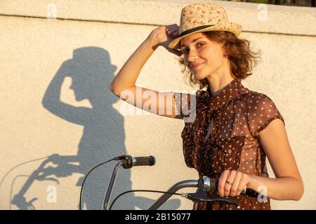 Junge hübsche Frau in Hut und Kleid mit Fahrrad und Schatten an der Wand mit Blick auf die Kamera lächelnd Stockfoto