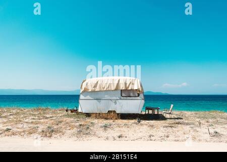 Reisemanhänger im Vintage-Stil an einem schönen Strand geparkt. Heller warmer Sommertag neben klarem türkisfarbenem Meerwasser. Hintergrund für den Urlaub Stockfoto