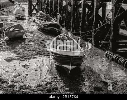 Boote sind im Schlamm in der Nähe eines Holzstegs mit ihren Festseilen noch befestigt. Die Holzstacheln des Stegs sind mit Barnacles verkrustet. Stockfoto