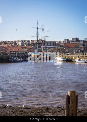Whitby Hafen an einem sonnigen Tag. Boote werden mit dem Nachbau der "HMS Endeavour" zu einem Wharf along vermauert. Ein Beitrag steht im Vordergrund. Stockfoto