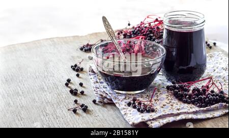 Hausgemachter schwarzer Elderberry-Sirup in Glasschale und -Glas und bunte schwarze Elderbeere im Hintergrund. Kopierbereich Stockfoto