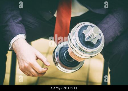 Geschäftsmann in schwarzem Anzug sitzt auf dem Stuhl und hält in der rechten Hand eine silberne Hanteln Stockfoto