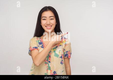 Hier werben! Faszinierendes asiatisches Mädchen mit brünettem Haar in der Sommer-Bluse, die beiseite zeigt und lächelt, Kopienraum zeigt, leerer Platz für Text Stockfoto