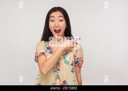 Sehen Sie, tolle Werbung hier! Faszinierendes asiatisches Mädchen mit brünettem Haar in der Sommer-Bluse, die beiseite zeigt und mit schockierter Ausdrucksweise aussieht und c zeigt Stockfoto