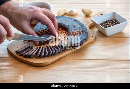 Man Hände schneiden Scheiben von spezieller Pirot-Hufeisenwurst - Peglana Pirotska kobasica, hergestellt aus Kalbfleisch, Schaf- und Ziegenfleisch Stockfoto