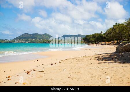 Sand, blaues Meer, Menschen und Hügel in der Ferne, Schatten von Mandelbäumen und der wolkige azurblaue Himmel geworfen Stockfoto