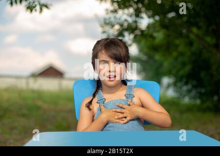 Witzig überraschte kleine Mädchen in einer Jeans-Sundress, zwei Schwänze, sitzt an einem Tisch im Sommergarten gegen einen blauen Himmel und grüne Blätter eines Baumabows Stockfoto