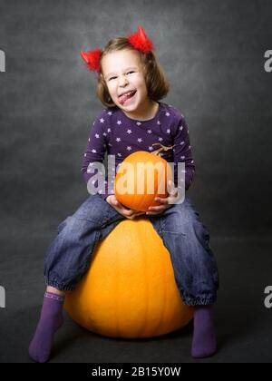 Halloween-Konzept. Kleines Mädchen mit Kostümhörnern, das Spaß hat. Adliges Kind sitzt auf Kürbis und Grimassen. Kleinkind lächelt und zeigt Zunge. Studio por Stockfoto
