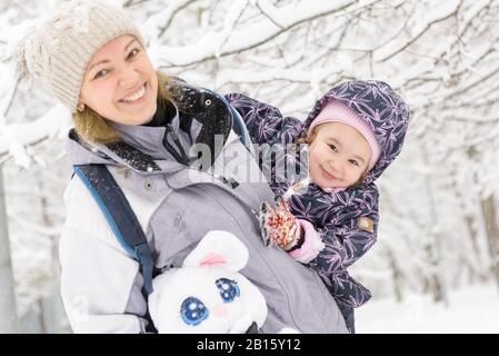 Süßes fröhliches kleines Kind spielt im Winter mit der Mutter in einem verschneiten Park. Fröhliche Familienspaziergänge bei Schneefall. Stockfoto