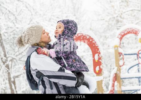 Fröhliches kleines Kind spielt im Winter mit der Mutter in einem verschneiten Park. Fröhliche Familienspaziergänge bei Schneefall. Süßes Baby-Mädchen sitzt auf den Händen der Mutter. Stockfoto