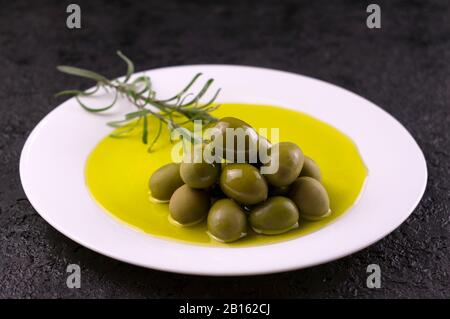 Oliven werden mit Olivenöl und einem Rosmarinspring auf einem weißen Teller gegossen. Das Konzept des Olivenöls. Stockfoto