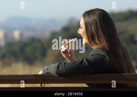 Eine glückliche Geschäftsfrau, die einen Snak auf einer Bank isst, während sie die Aussicht im Freien beobachtet Stockfoto