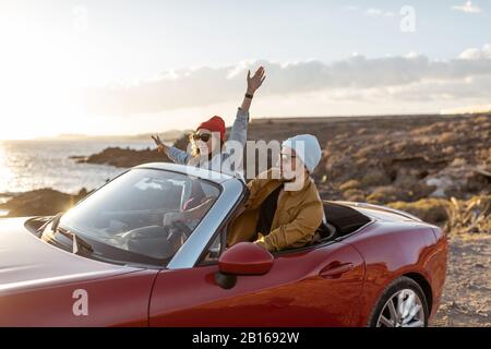 Fröhliches Paar genießt Ferien und fährt zusammen ein Cabrio-Auto an der felsigen Küste des Ozeans bei Sonnenuntergang. Glücklicher Urlaub, Liebe und Reisekonzept Stockfoto