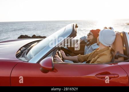 Fröhliches Paar genießt Ferien und fährt zusammen ein Cabrio-Auto an der felsigen Küste des Ozeans bei Sonnenuntergang. Glücklicher Urlaub, Liebe und Reisekonzept Stockfoto