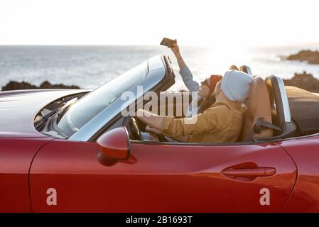 Fröhliches Paar genießt Ferien und fährt zusammen ein Cabrio-Auto an der felsigen Küste des Ozeans bei Sonnenuntergang. Glücklicher Urlaub, Liebe und Reisekonzept Stockfoto