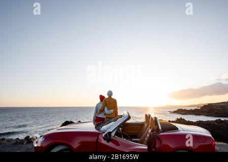 Ein Paar, das einen schönen Blick auf den Ozean genießt und sich in der Nähe des Autos an der felsigen Küste umarmt, einen weiten Blick von der Seite mit Platz am Himmel Stockfoto