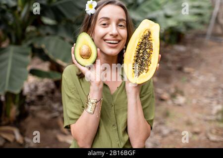 Porträt einer jungen lächelnden Frau mit geschnittener Papaya-Frucht und Avocado-Beere im Freien auf der tropischen Plantage. Konzept von Vegetarismus, gesundem Essen und Wohlbefinden Stockfoto