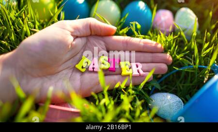 Frohe Ostern Banner. Ostereier versteckt im Frühlingsgras. Schreiben aus Holzbuchstaben in der Hand. Grußkarte. Frühling Stockfoto