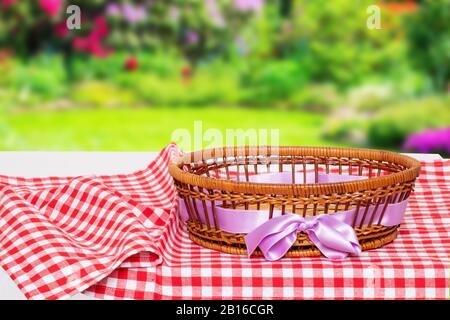 Tischplatte auf leerem Korbhintergrund. Leerer Picknickkorb mit rosafarbenem Band auf rotem kariertem Tischtuch mit natürlichem, verschwommenem Sommergarten-Hintergrund. Stockfoto