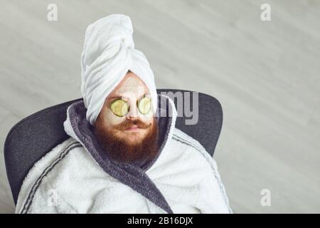 Lustiger fetter bärtiger Mann mit einer kosmetischen Maske im Gesicht in einem bademantel auf dem Kopf auf dem Gesicht, der vor einem grauen Hintergrund ruht Stockfoto