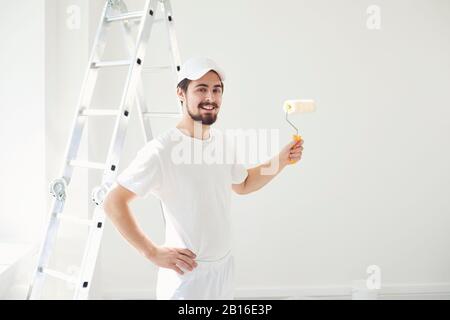 Ein männlicher Maler in weißer Uniform mit einer Walze arbeitet in der Hand in einem weißen Raum Stockfoto